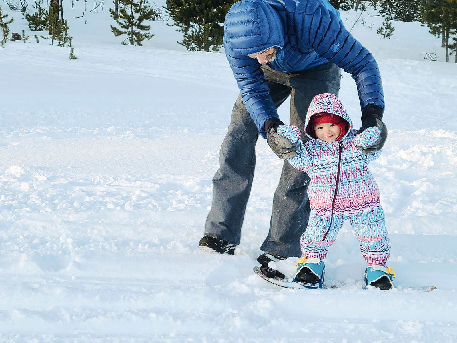 A dad helping his 1-year-old baby get used to gliding on a snowboard