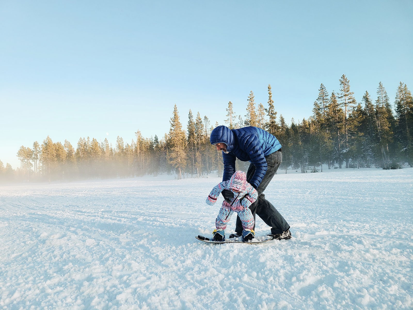 Toddler flopping like a ragdoll on her snowboard as her dad holds her from behind