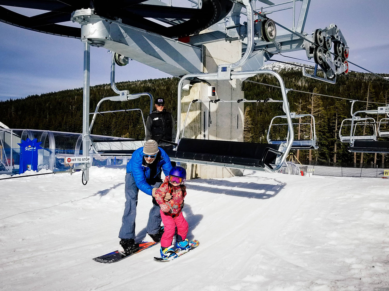 A dad helping his 3-year-old toddler get off the chairlift on snowboards