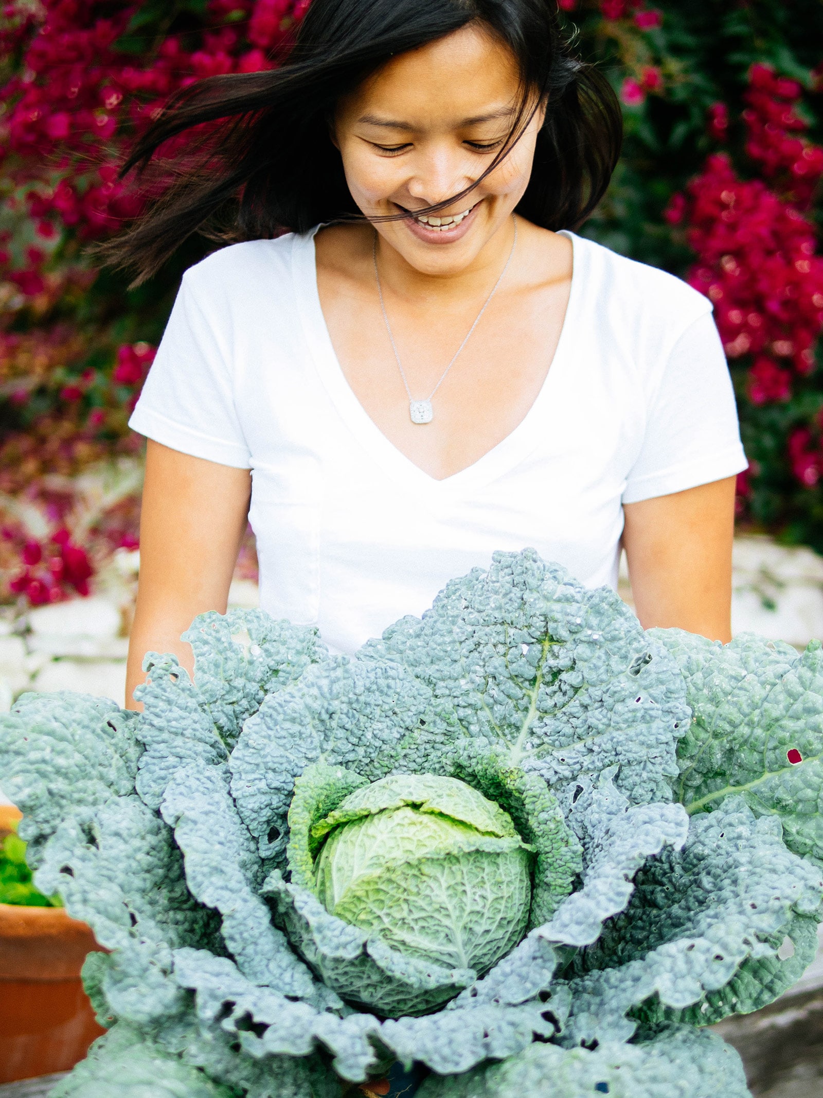 Woman (Linda Ly) holding a large head of cabbage in a garden, looking down at it and smiling