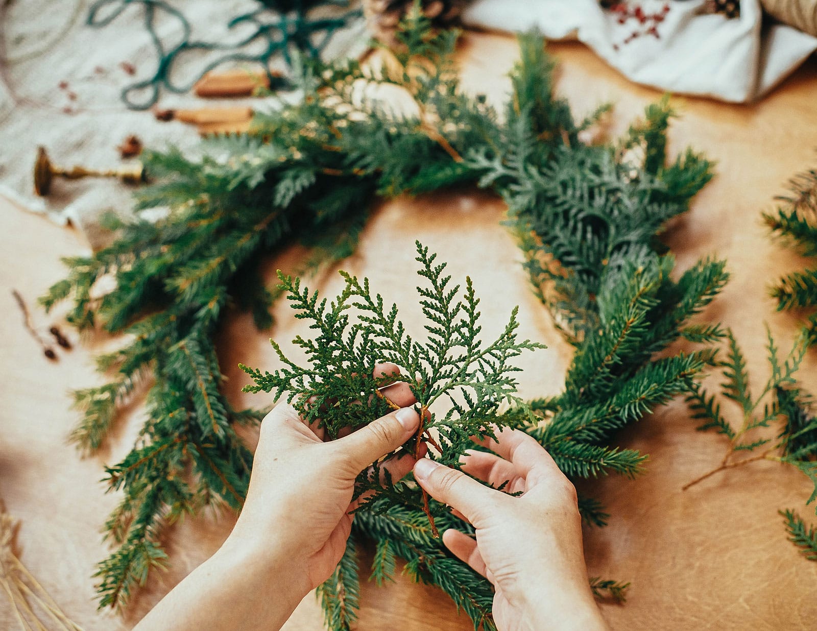 Hands holding a fresh-cut evergreen branch with a homemade wreath in the background