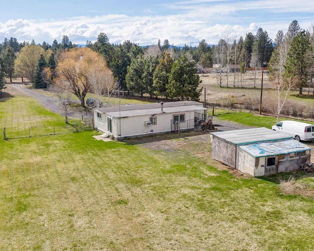 Real estate listing photo showing a rural grassy property with an old manufactured home and rustic wooden sheds