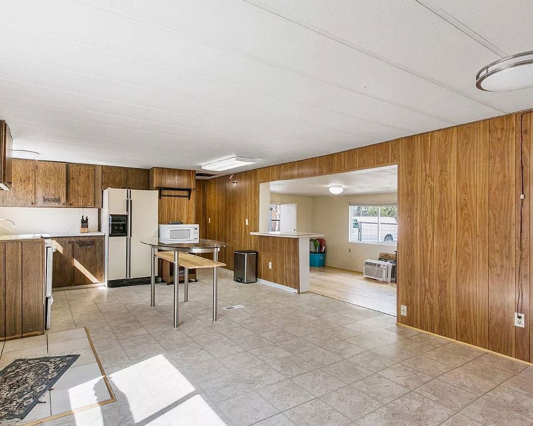 Real estate listing photo showing the interior of an older manufactured home with faux wood paneling, vinyl floors, and ceiling tiles