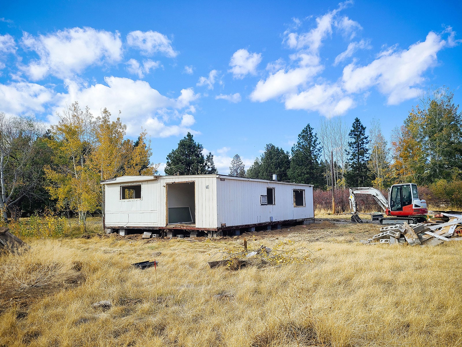 Older white manufactured home with missing windows and doors in a field of yellow grass