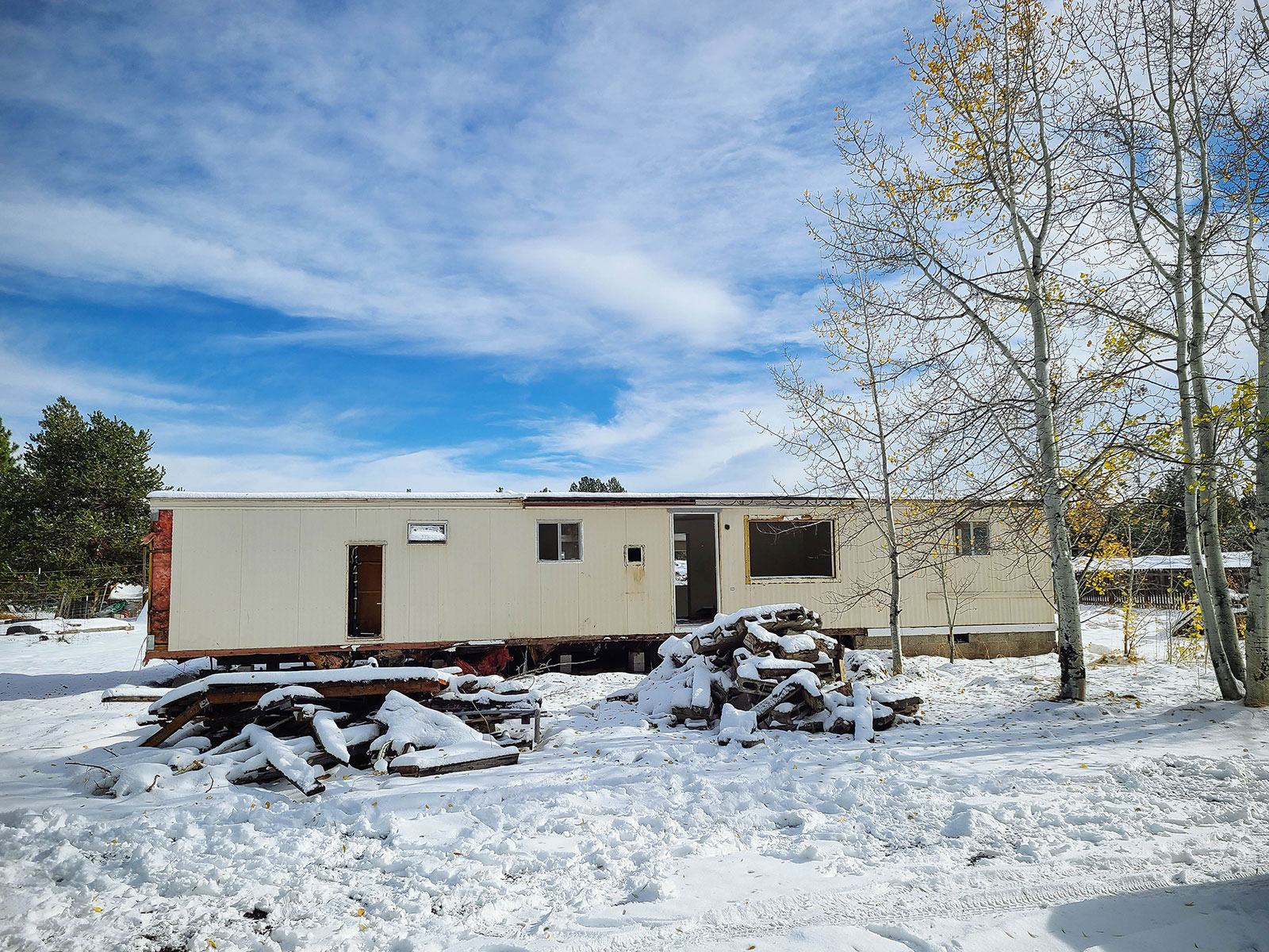 Older manufactured home with missing windows and doors, set in a snowy yard with debris piles in front