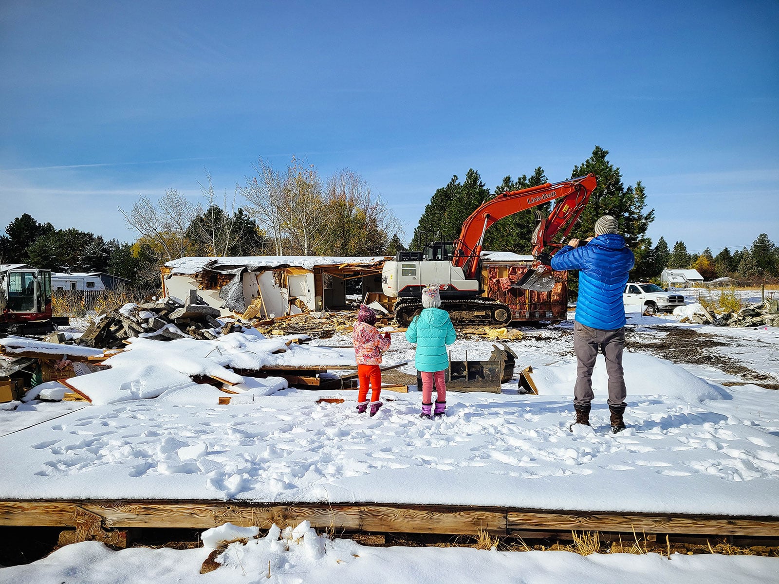 Man in blue jacket and two daughters watching a small house get demolished by an excavator in the snow