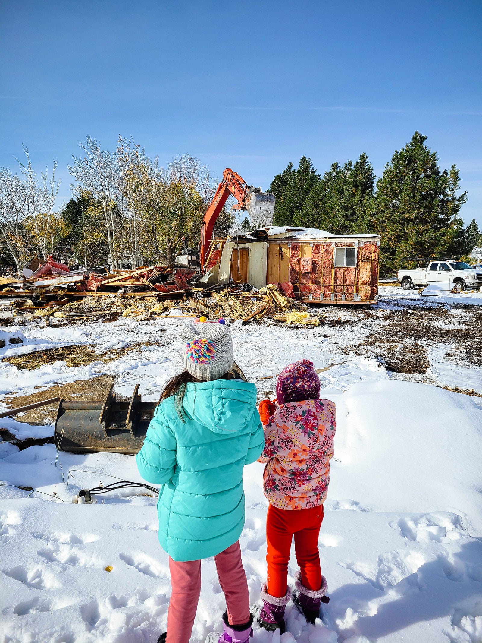 Two children in winter clothing watching a house get demolished in the snow
