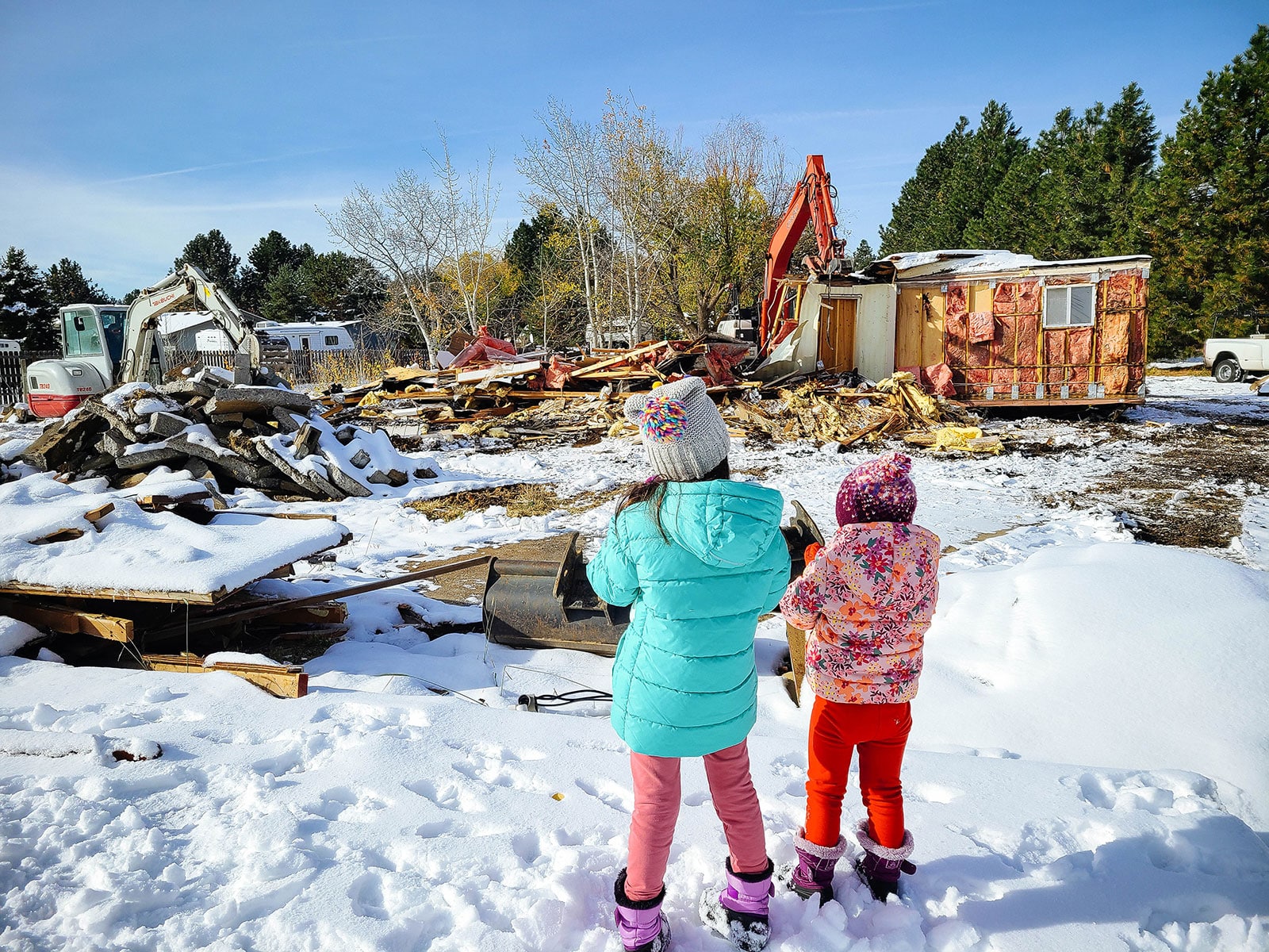 Two children watching a house get demolished by an excavator in a snowy yard