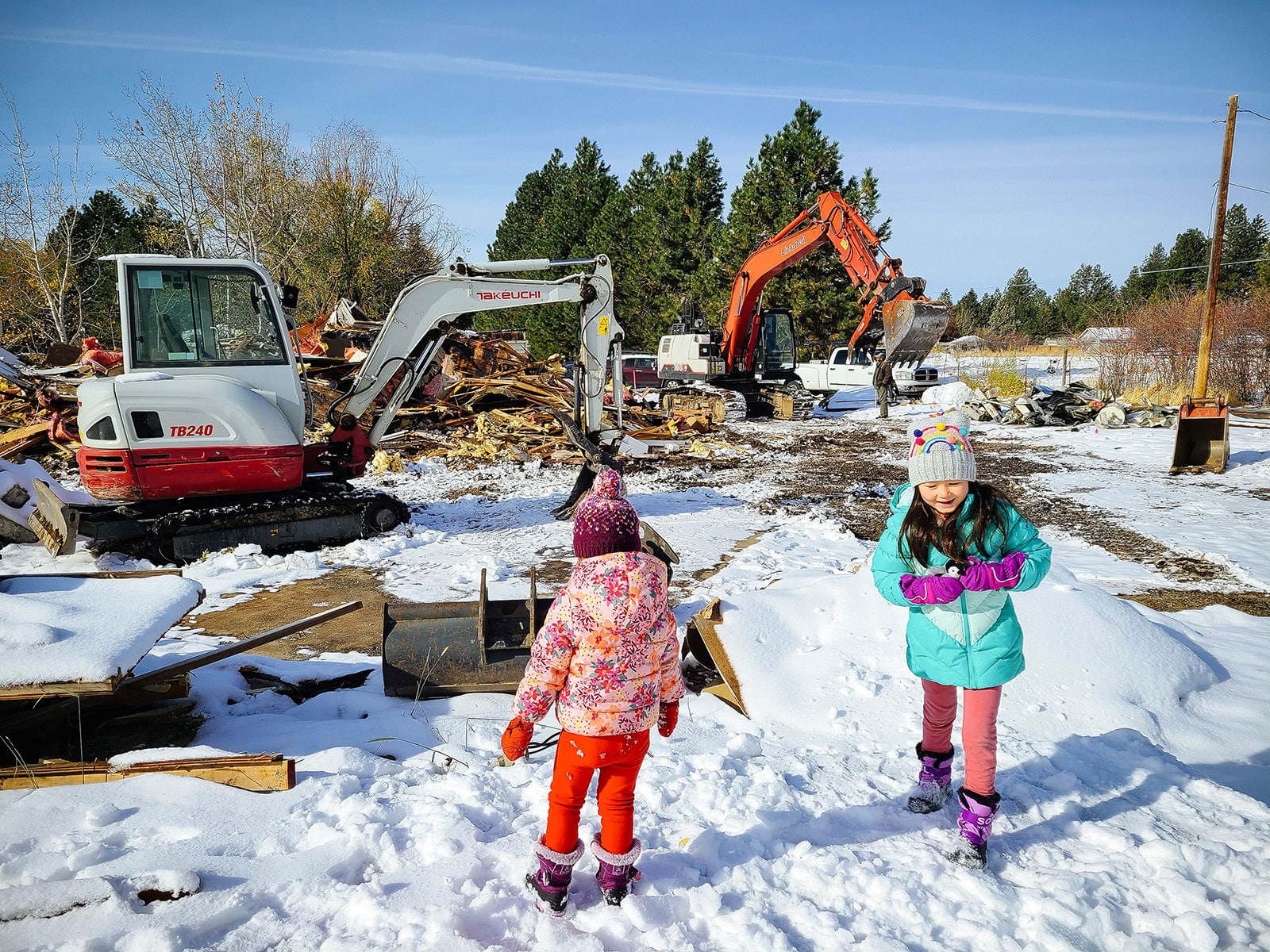 Two children watching excavators clean up debris piles from a snow-covered property