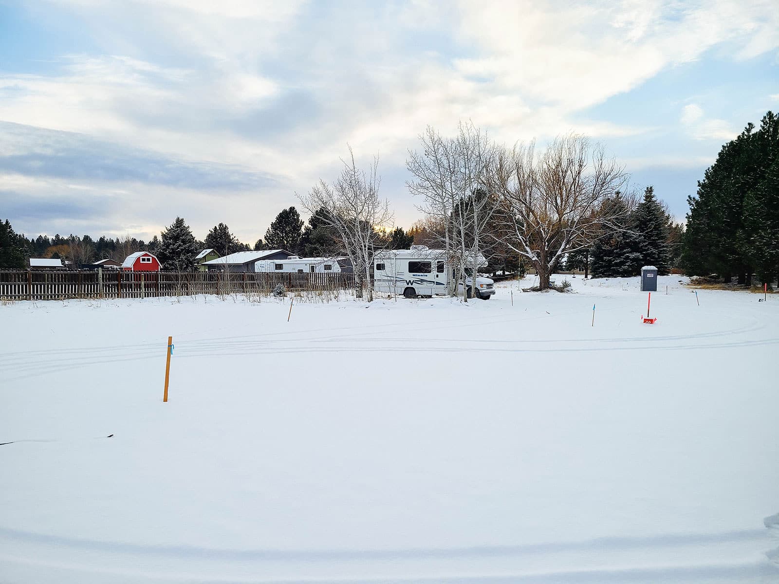 A snowy yard with survey stakes in the ground and an RV parked in the background