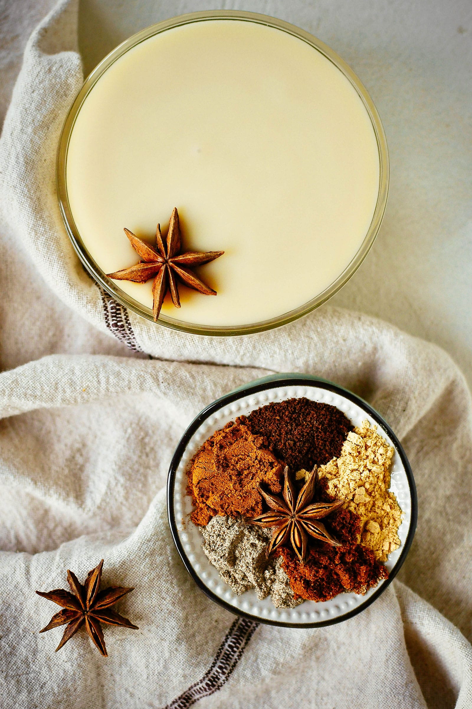 A glass bowl of condensed milk next to a small dish filled with ground chai spices, shot against a linen napkin background