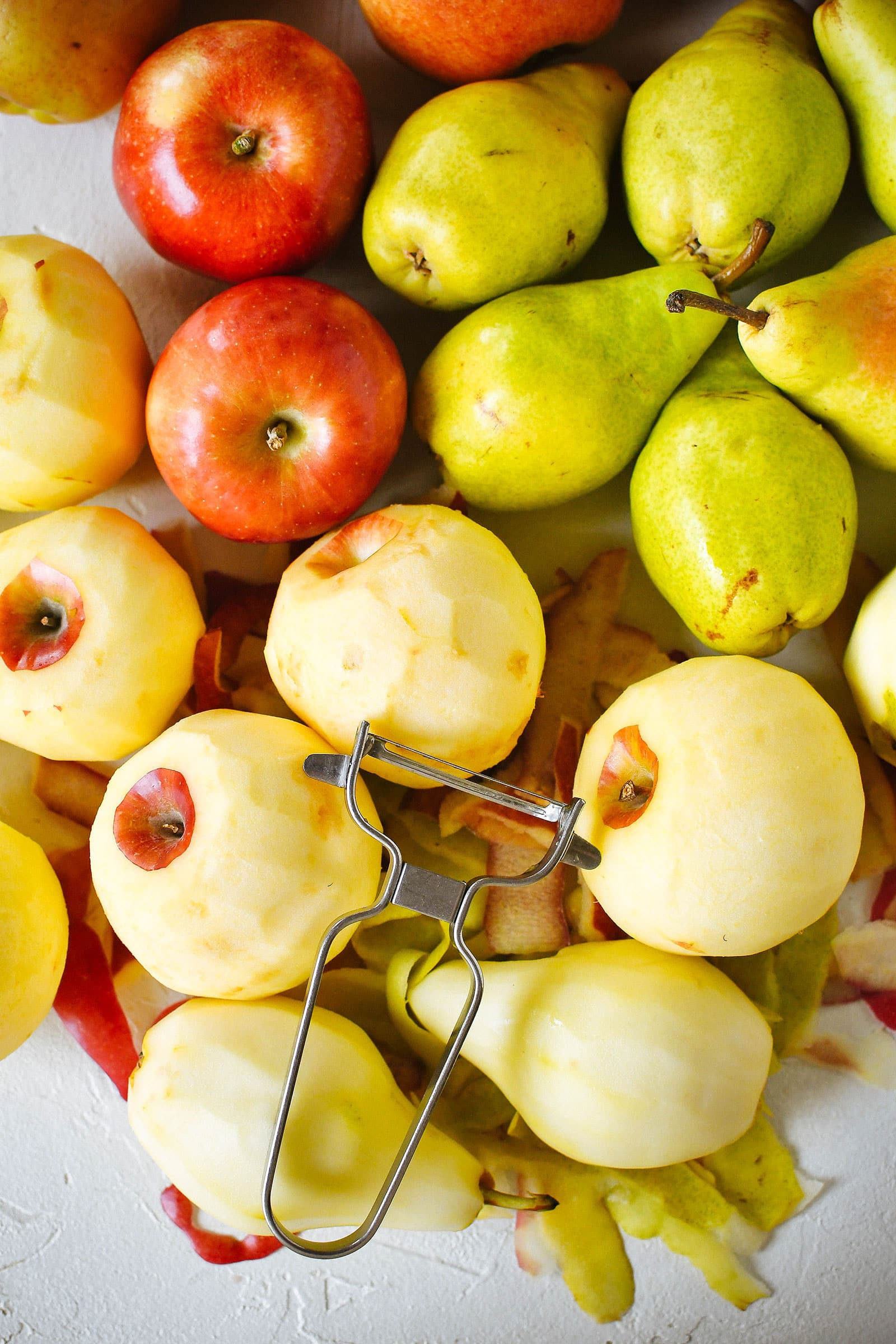 A pile of red apples and green pears, with some fruits peeled next to a metal peeler
