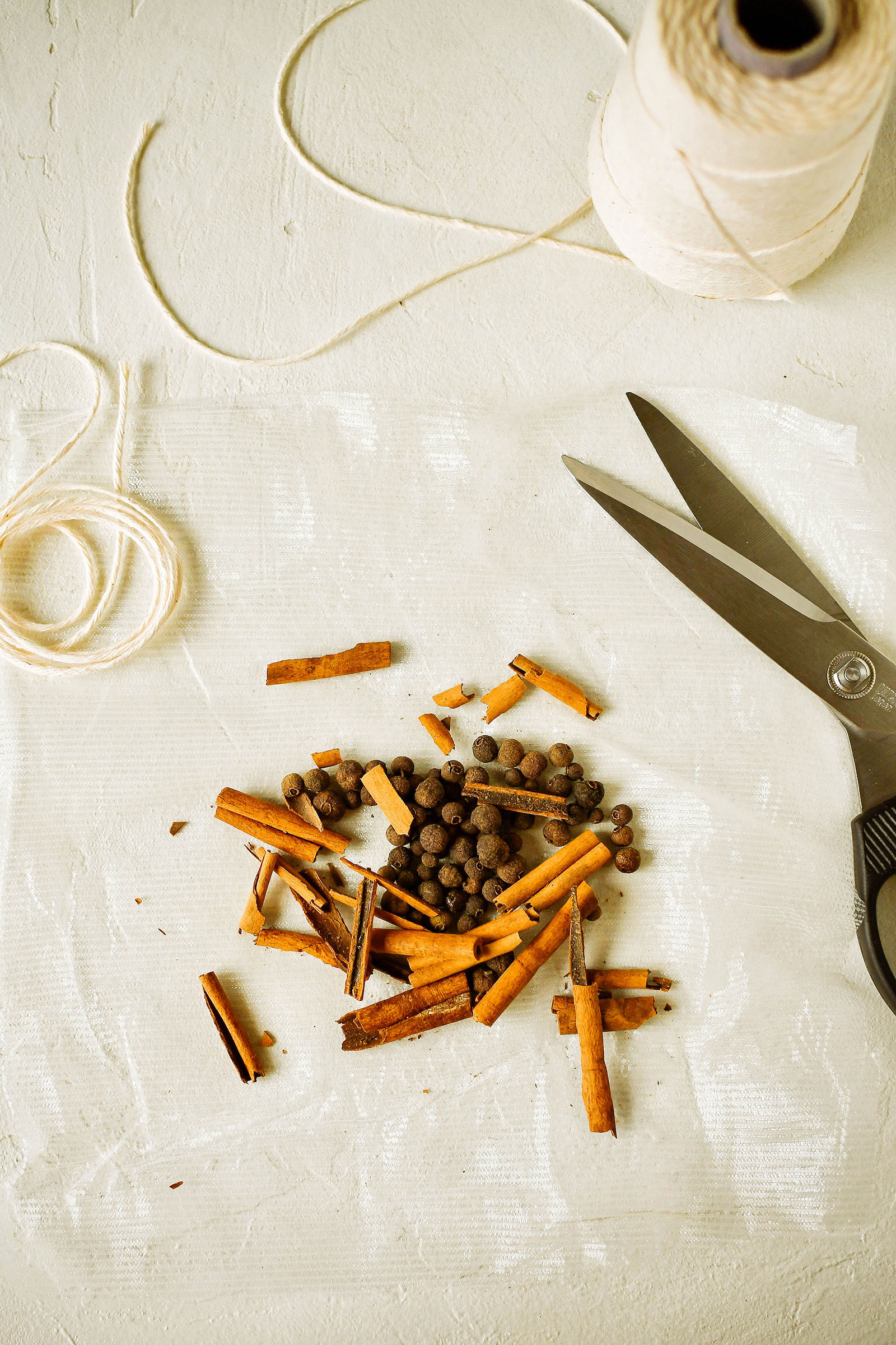 Allspice berries and broken cinnamon sticks on a white film, next to a roll of twine and a pair of scissors