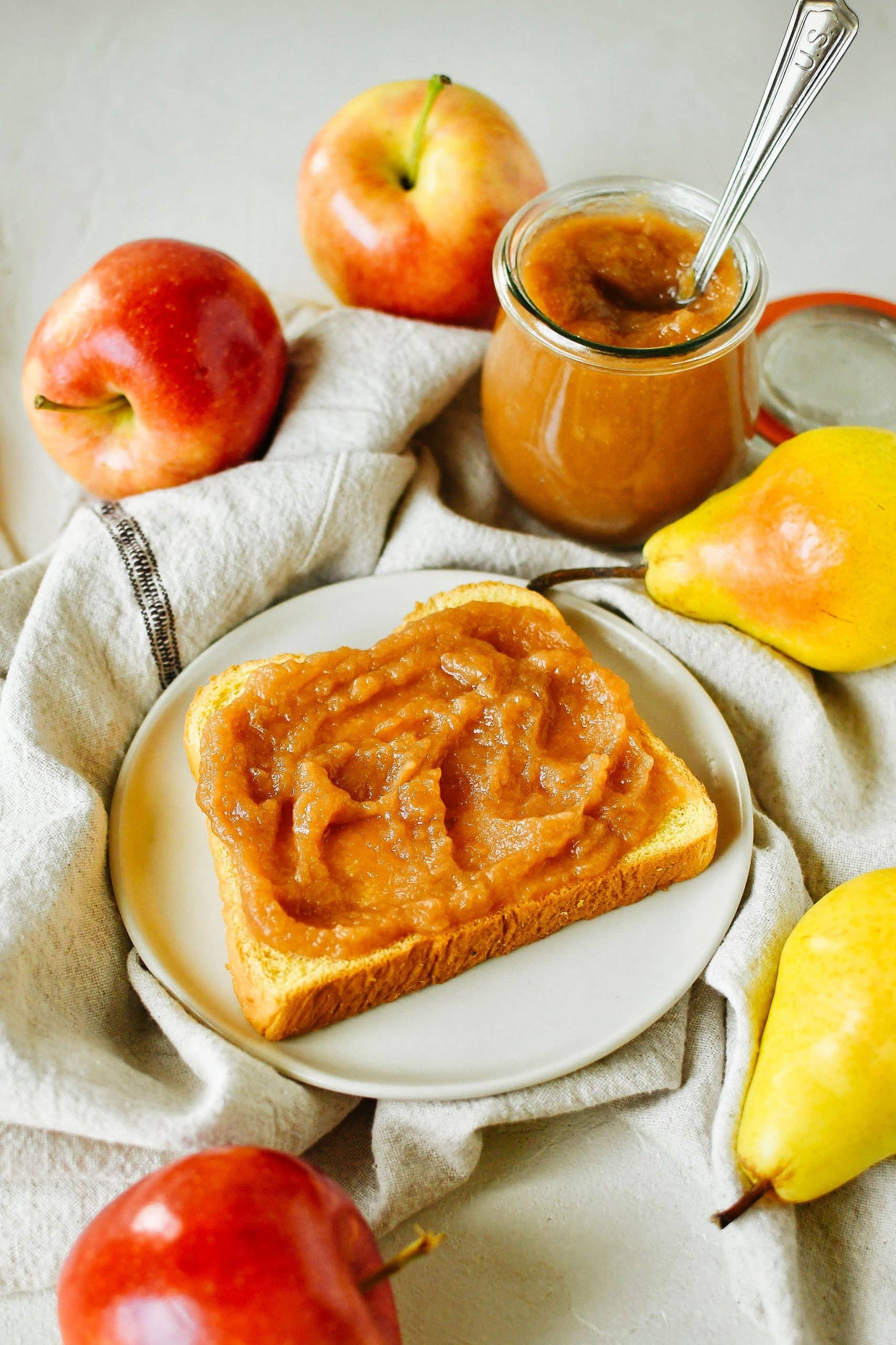 Piece of toast on a white plate, smothered in apple-pear butter, with apple and pear fruits arranged around the plate next to a jar of apple-pear butter