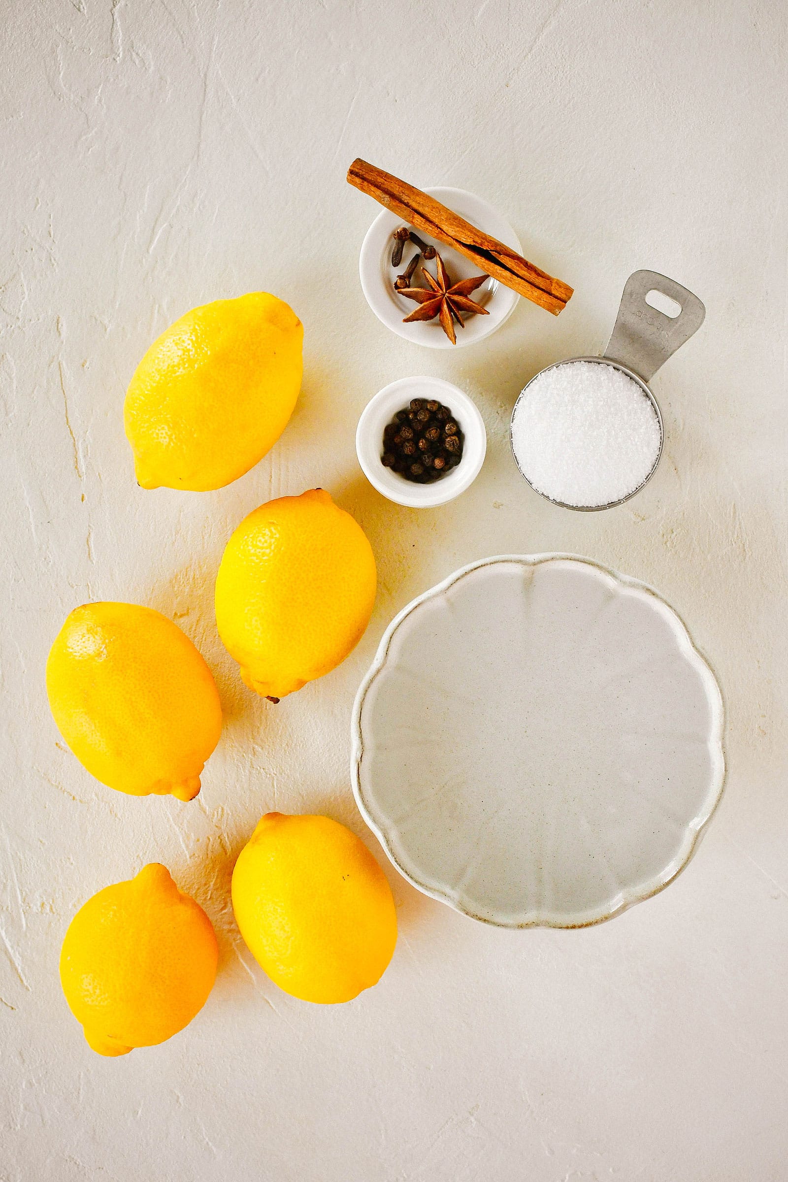 Overhead shot of a bowl of water, a measuring cup filled with salt, small dishes with various whole spices, and five lemons on a table