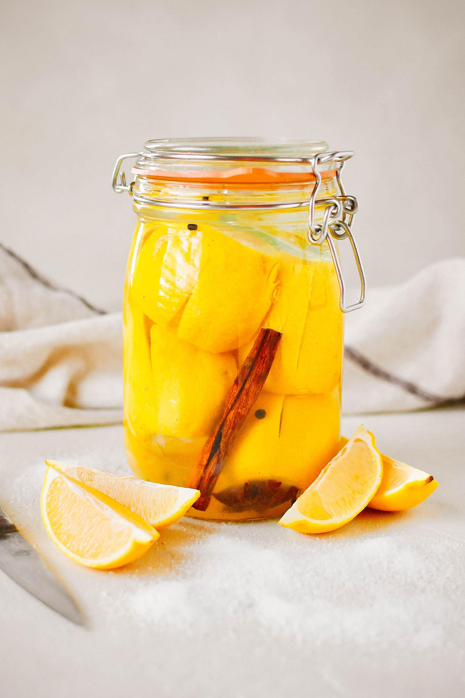 Stylized shot of a jar of Moroccan preserved lemons with cut lemons arranged around it and a linen napkin in the background