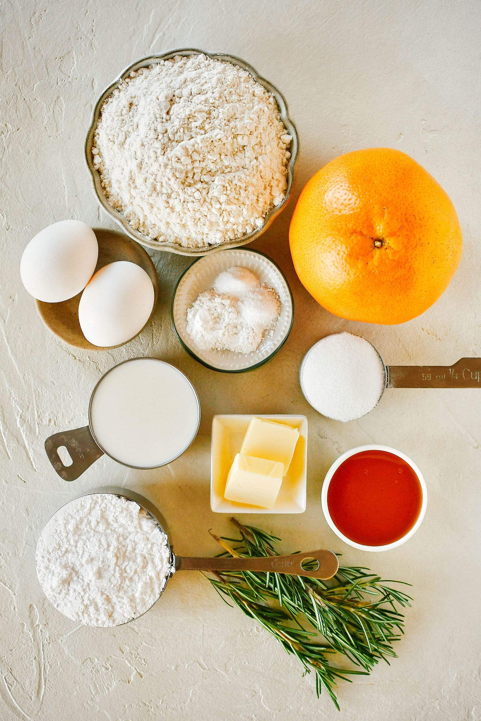 Overhead shot of grapefruit-rosemary bread ingredients on a white surface, including flour, sugar, eggs, butter, buttermilk, baking soda, salt, honey, grapefruit, and rosemary sprigs