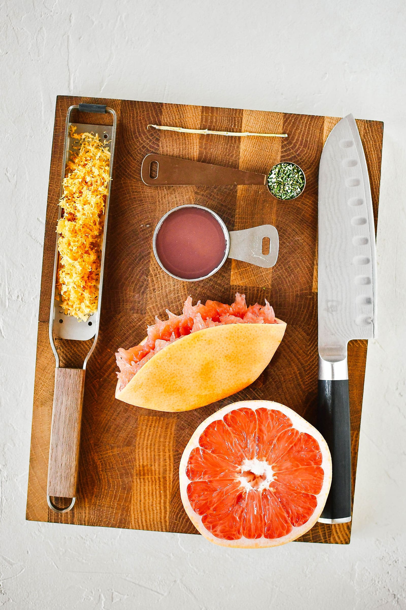 Overhead shot of bread ingredients laid out on a wooden cutting board, including a zested grapefruit, juiced grapefruit, and minced rosemary with a knife next to them