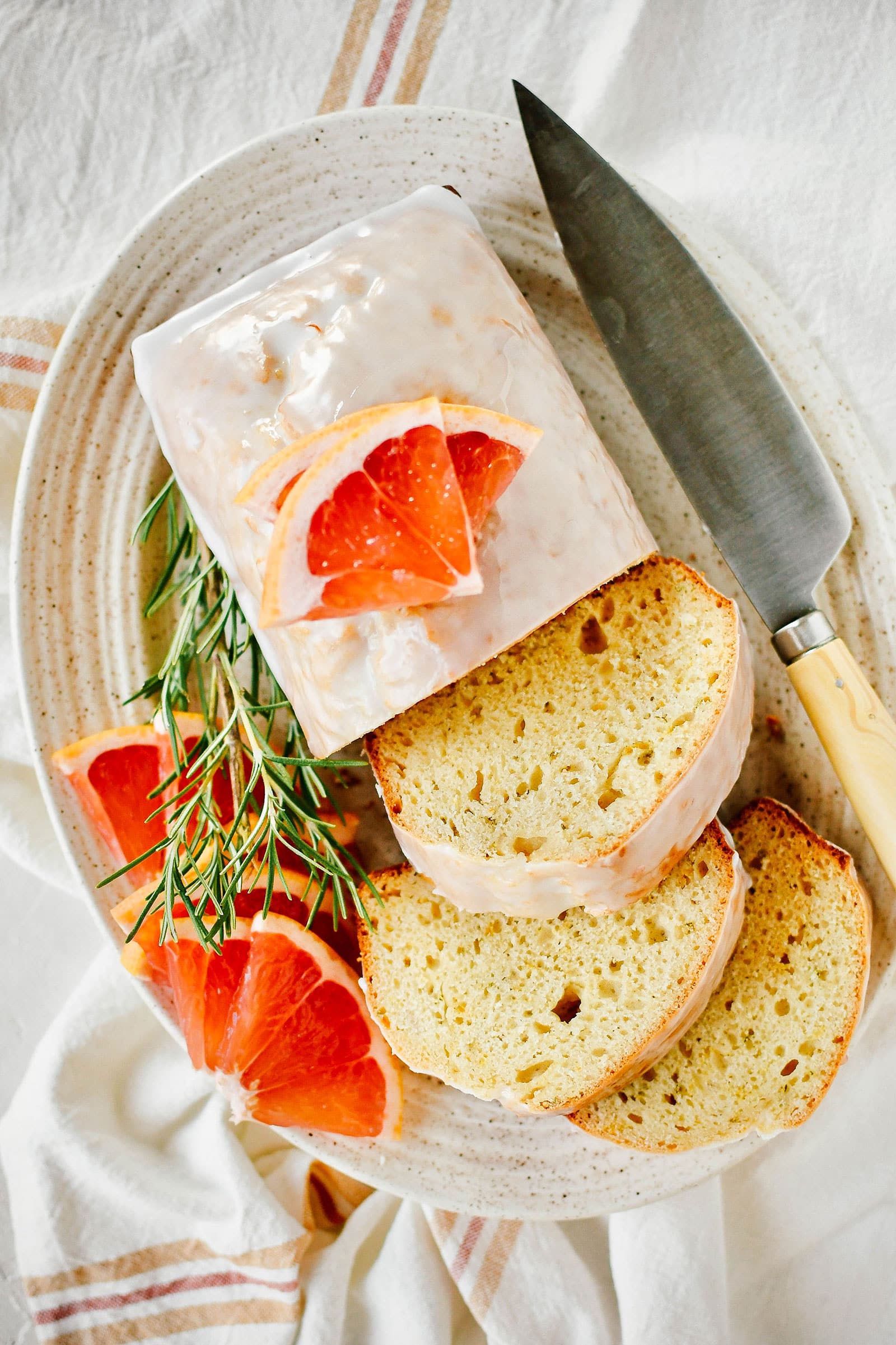 Stylized overhead shot of grapefruit-rosemary bread on a speckled ceramic platter, sliced open with a knife next to the loaf