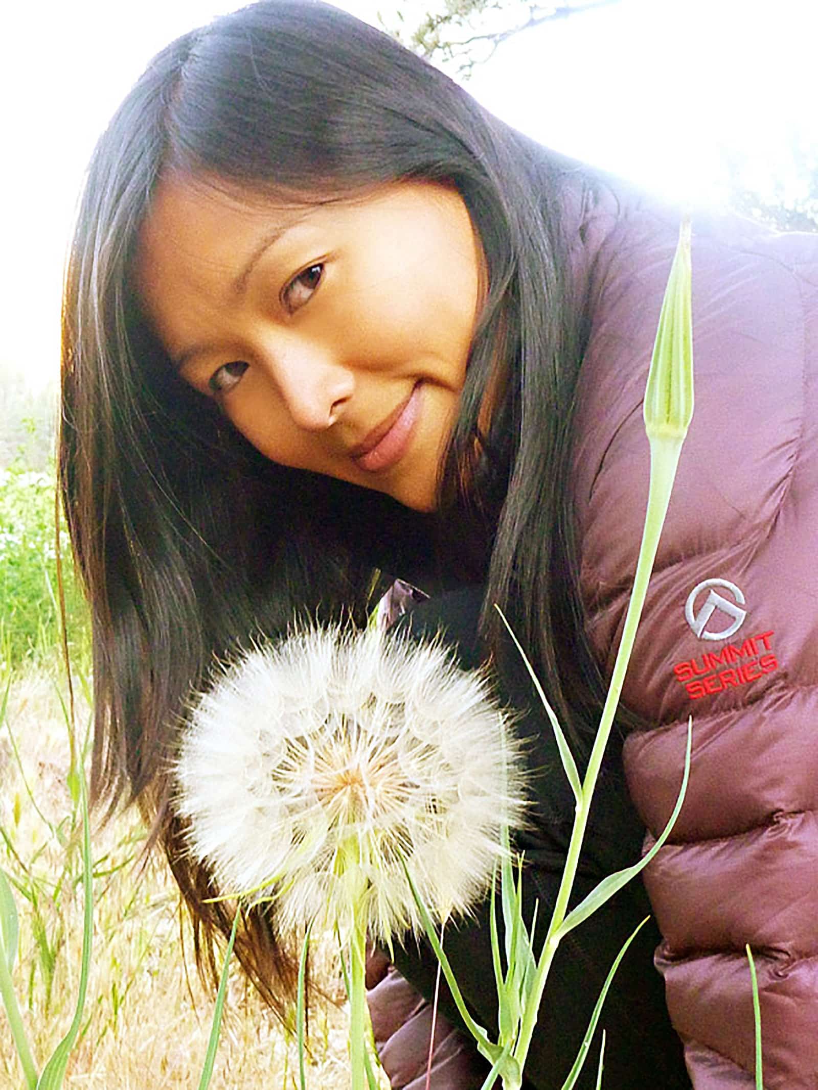 Linda Ly in a maroon puffy jacket, sitting behind a large salsify flower seed head in a meadow