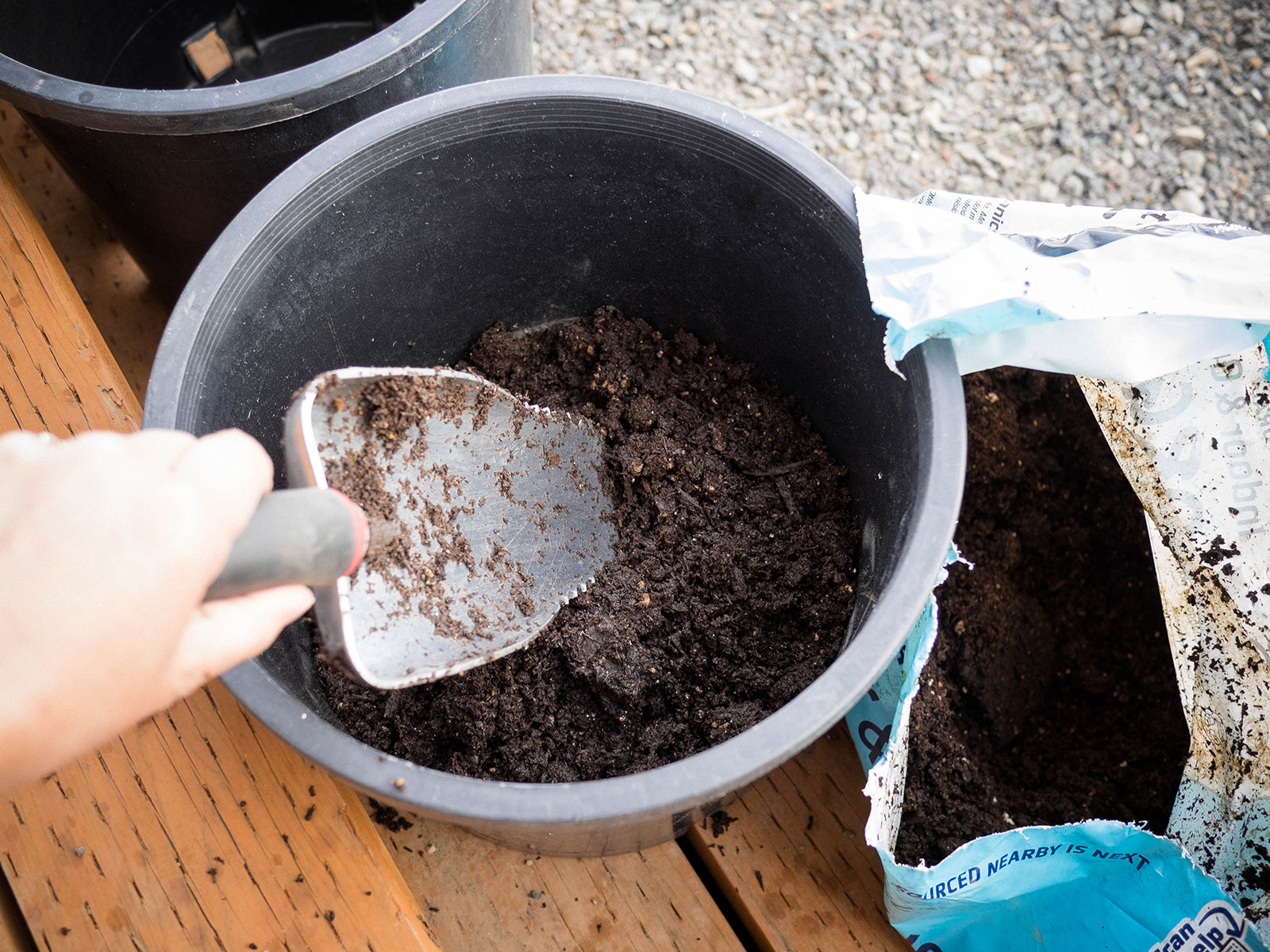Hand holding a trowel and filling a large black plastic nursery pot with potting soil