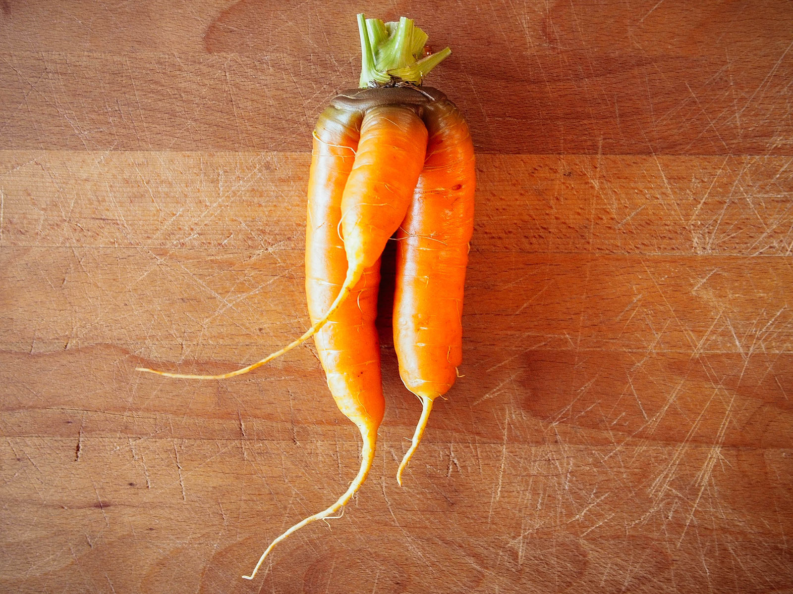 An orange carrot on a wooden cutting board with two long legs and a shorter, stout leg between them