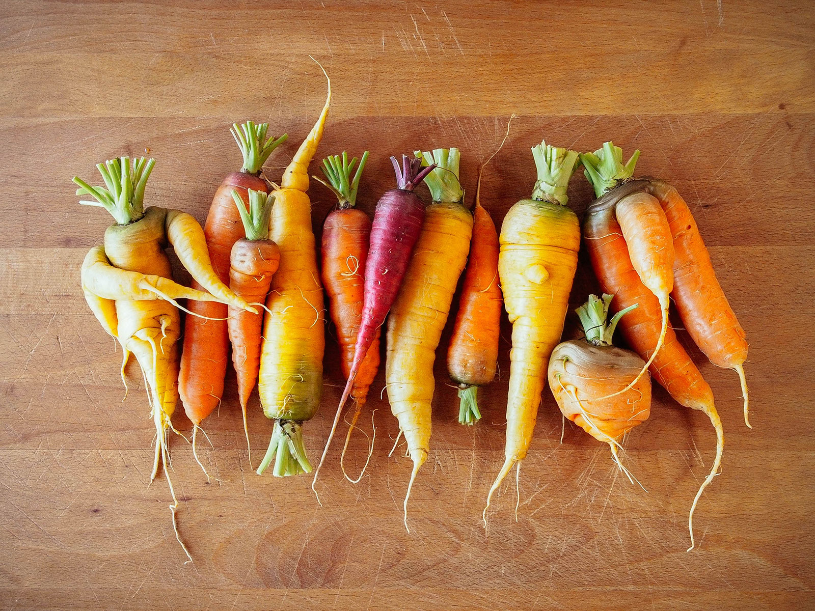 A group of colorful yellow, orange, and purple carrots of all shapes and sizes (some with forked roots) on a wooden cutting board