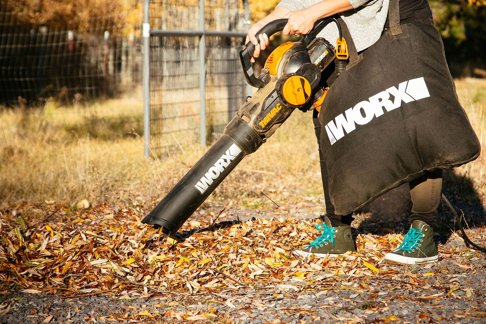 Woman in a yard, using a leaf blower with a mulching blade and bag attachment to vacuum and shred a small mound of fall leaves