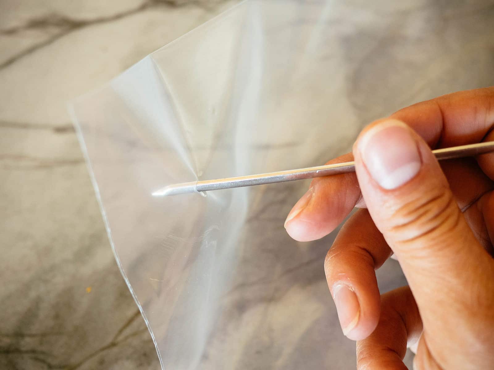 A hand using a metal skewer to poke small holes through a plastic zip-top bag