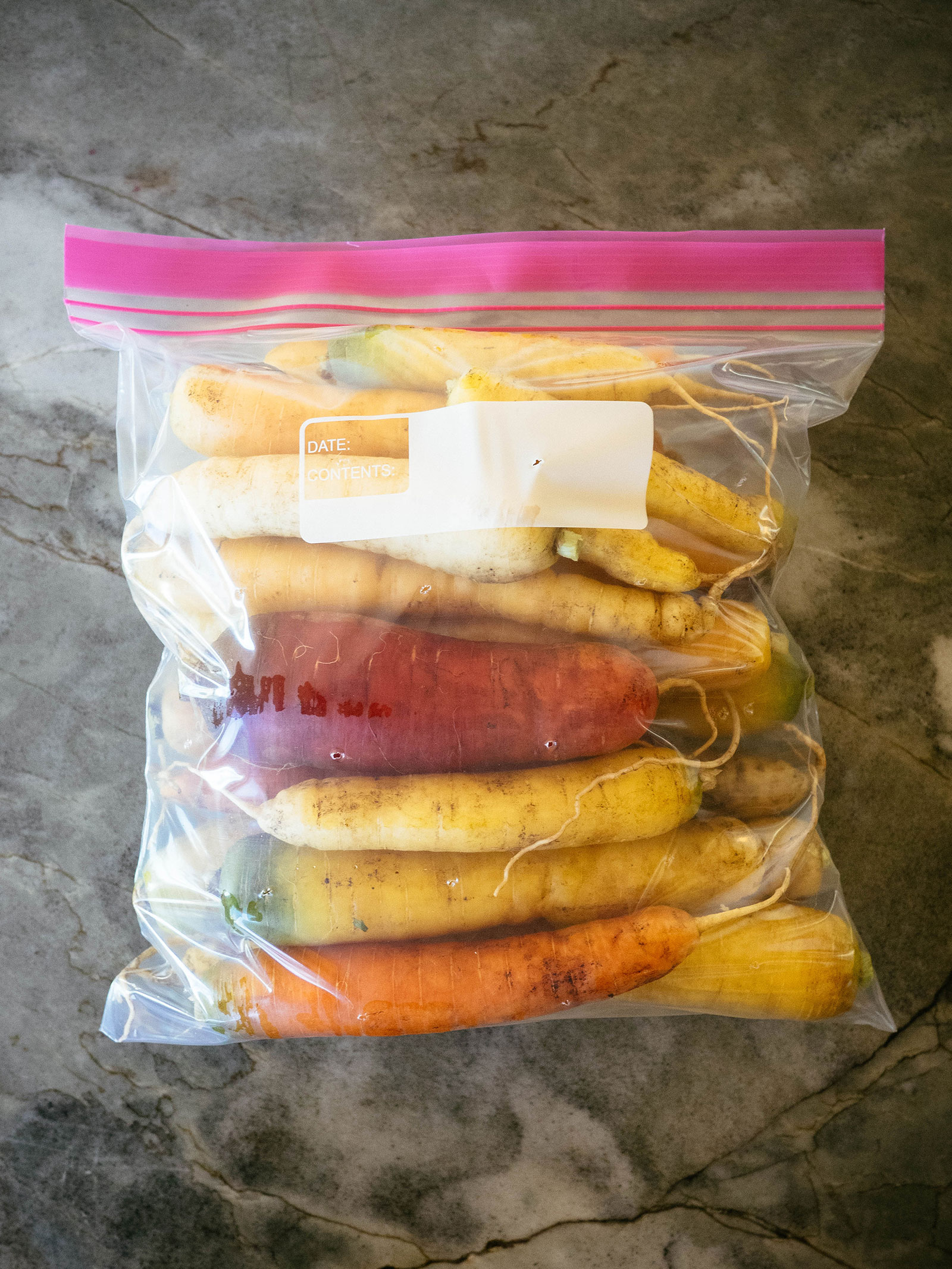 A gallon-size zip-top bag on a stone countertop, filled with colorful carrots from the garden