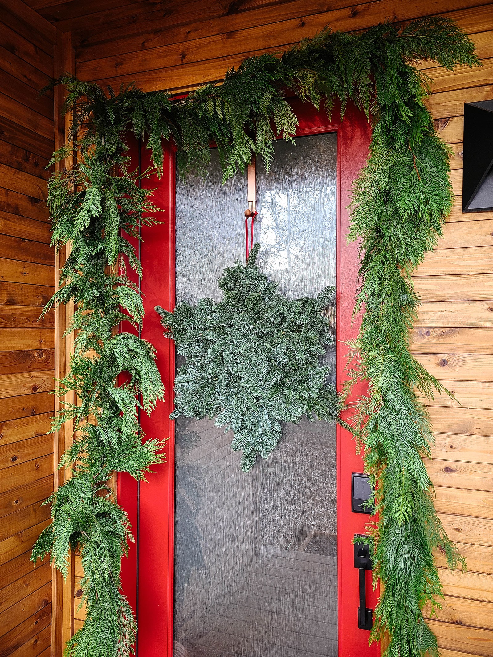 A snowflake wreath and cedar garland decorating a glass-paneled front door on a wood-encased porch