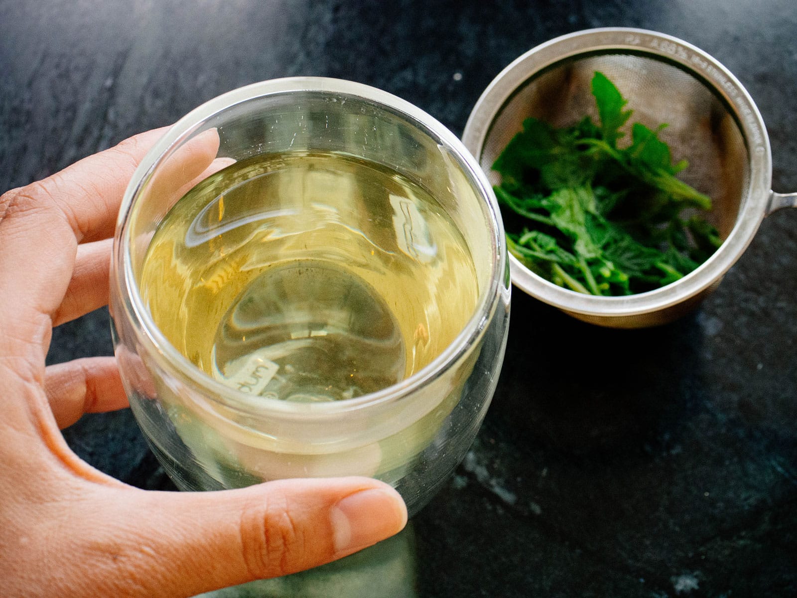 Hand holding a glass cup filled with tomato leaf tea, with a strainer of steeped tomato leaves on the side