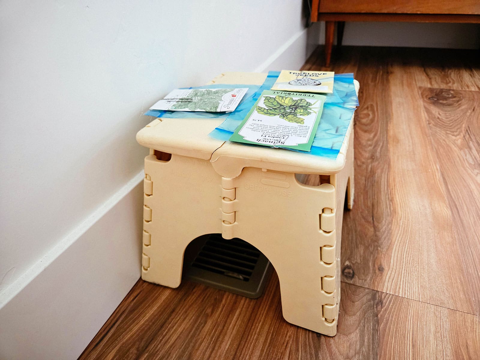 Packets of seeds clipped into small blue bags holding spinach seeds sit on a beige stool above a floor vent